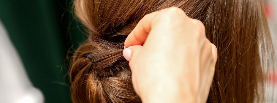 Hands of hairdresser making french twist hairstyle of an unrecognizable young brunette woman in a beauty salon, back view, close up