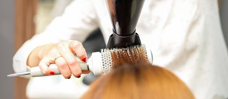 A professional hairdresser is drying long red hair with a hair dryer and round brush, close up