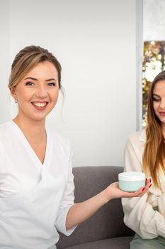 Beautician offering product for young woman, shows thumb up, holding a white plastic jar with a cream, mockup, copy space