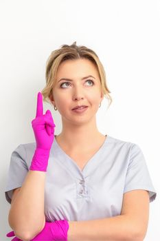 Portrait of young caucasian doctor woman wearing rubber gloves in medical uniform with one finger up, looking up against a white background, copy space