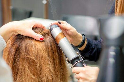 Hands of female hairstylist curls hair client with a curling iron in a hairdressing salon, close up