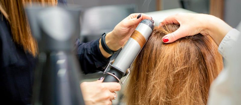 Hands of female hairstylist curls hair client with a curling iron in a hairdressing salon, close up