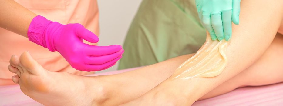 Sugaring legs. Woman legs hair removing. Hands in pink and blue rubber gloves of two beauticians apply sugar paste on female feet