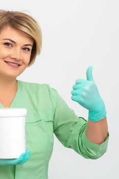 Beautician in gloves holding a jar of the cosmetic product showing thumb up on a white background