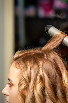 Hairdresser makes curls with a curling iron for the young woman with long brown hair in a beauty salon