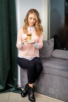 Beautiful caucasian young woman with long wavy blonde hair holding and drinking latte from a glass cup, sitting on a sofa