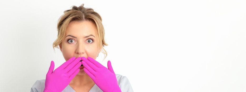 Portrait of a young female caucasian doctor or nurse is shocked covering her mouth with her pink gloved hands against a white background