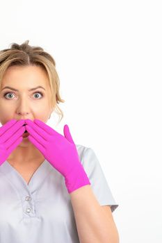 Portrait of a young female caucasian doctor or nurse is shocked covering her mouth with her pink gloved hands against a white background