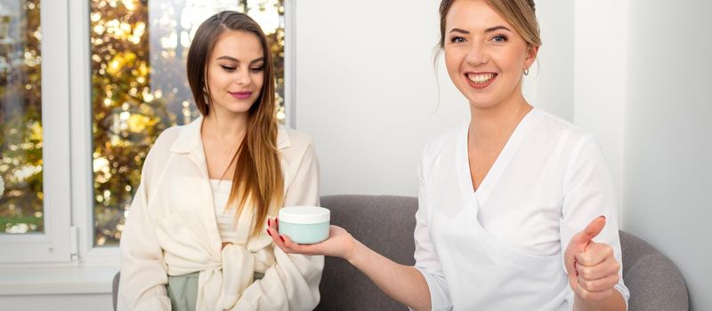 Beautician offering product for young woman, shows thumb up, holding a white plastic jar with a cream, mockup, copy space