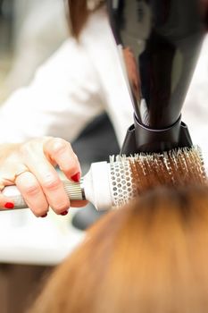 A professional hairdresser is drying long red hair with a hair dryer and round brush, close up