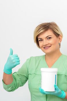 Beautician in gloves holding a jar of the cosmetic product showing thumb up on a white background