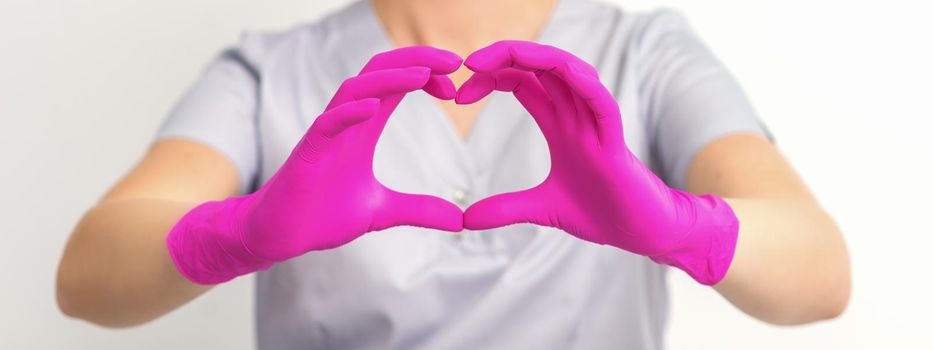 A smiling caucasian woman doctor wearing pink gloves in uniform showing the symbol of a heart against a white background