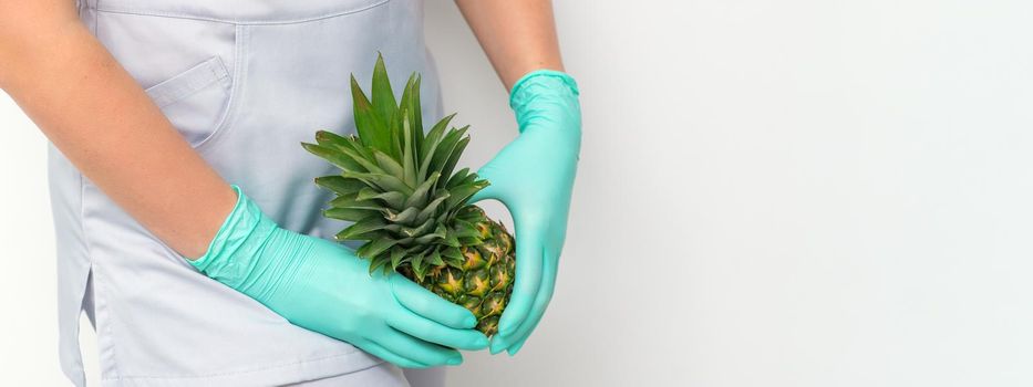 Young beautician wearing blue gloves in uniform with pineapple covers an intimate area on a white background, bikini zone depilation concept
