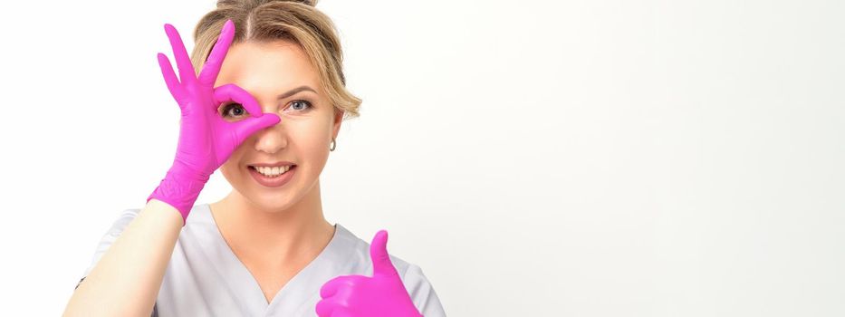 Smiling doctor oculist caucasian woman wearing pink rubber gloves in uniform showing ok sign covering the eye and thumb up gesture against a white background