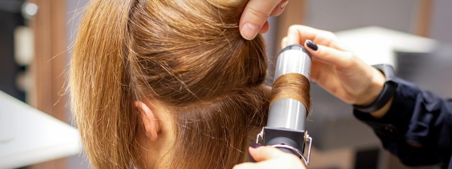 Hands of female hairstylist curls hair client with a curling iron in a hairdressing salon, close up