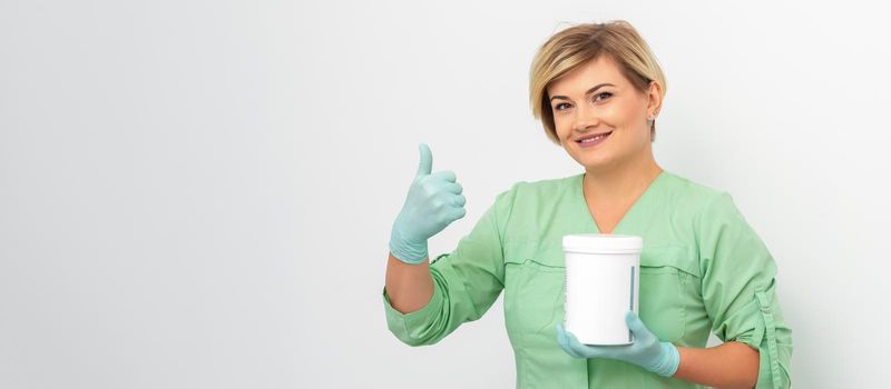 Beautician in gloves holding a jar of the cosmetic product showing thumb up on a white background