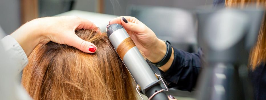 Hands of female hairstylist curls hair client with a curling iron in a hairdressing salon, close up
