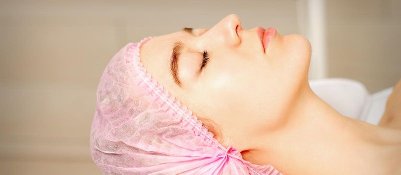 Side view of a young caucasian woman in a medical cap lying with closed eyes waiting for the cosmetic procedure in a beauty salon