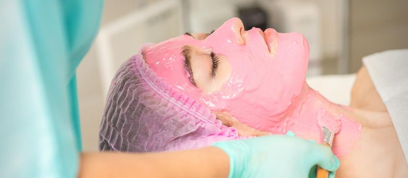 Hand of cosmetologist applying the pink alginic mask with the brush to the face of the young woman in a beauty salon