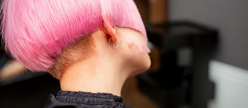 Styling and cutting dyed pink hair of young model back view in a hairdressing salon. Pink short hairstyle