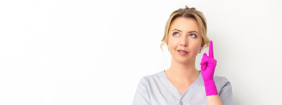 Portrait of young caucasian doctor woman wearing rubber gloves in medical uniform with one finger up, looking up against a white background, copy space