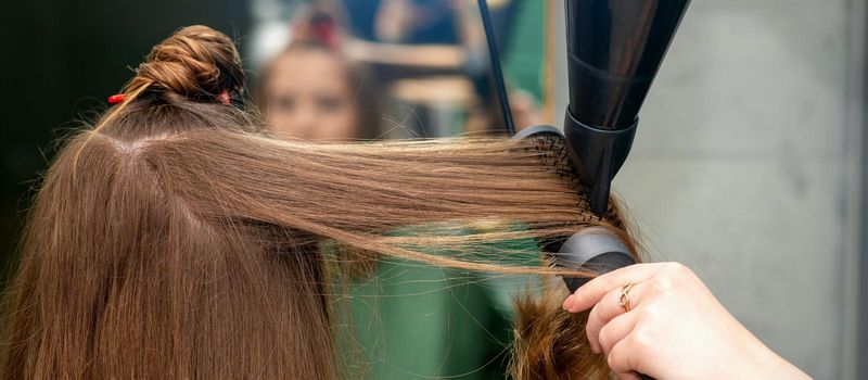 A hairdresser is drying long brown hair with a hairdryer and round brush in a hairdressing salon