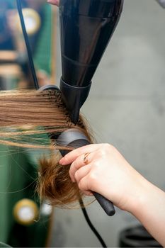 A hairdresser is drying long brown hair with a hairdryer and round brush in a hairdressing salon