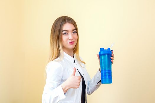 The young Caucasian female doctor wearing a white coat holds a blue plastic bottle with water with thumb up looking at the camera
