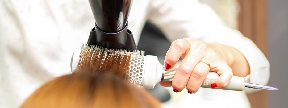 A professional hairdresser is drying long red hair with a hair dryer and round brush, close up