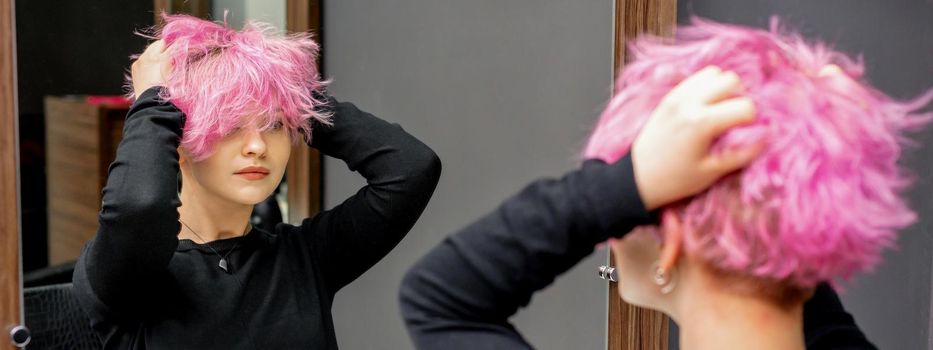 Young woman checking her new curly short pink hairstyle with he hands in front of the mirror at the hairdresser salon.