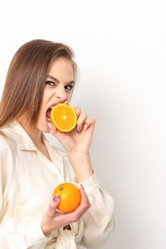 Young caucasian pretty cunning brunette woman biting one orange half and looking at the camera wearing a white shirt over white background