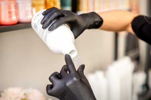 A hairdresser in black gloves is preparing hair dye with a bottle in a hair salon, close up