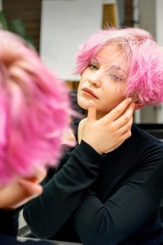 The beautiful young caucasian woman with a new short pink hairstyle looking at her reflection in the mirror checking hairstyle in a hairdresser salon