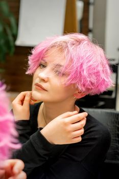 The beautiful young caucasian woman with a new short pink hairstyle looking at her reflection in the mirror checking hairstyle in a hairdresser salon