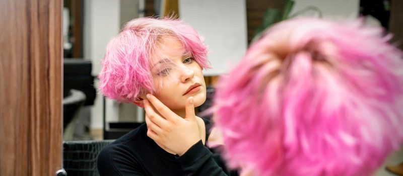 The beautiful young caucasian woman with a new short pink hairstyle looking at her reflection in the mirror checking hairstyle in a hairdresser salon