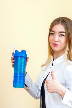 The young Caucasian female doctor wearing a white coat holds a blue plastic bottle with water with thumb up looking at the camera