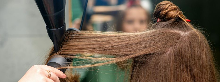 A hairdresser is drying long brown hair with a hairdryer and round brush in a hairdressing salon