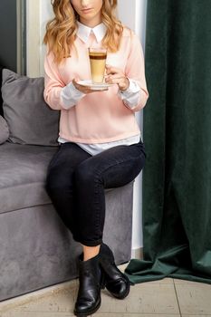 Beautiful caucasian young woman with long wavy blonde hair holding and drinking latte from a glass cup, sitting on a sofa