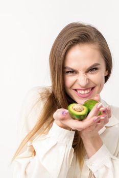 Portrait of a lovely smiling young brunette caucasian woman wearing the white shirt with long hair holding and showing avocado, standing isolated over white background