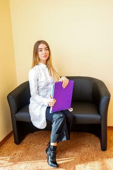 Portrait of a young caucasian doctor with folders and blonde long hair wearing a white coat sitting on the black sofa looking at the camera