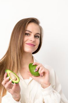 Portrait of a lovely smiling young brunette caucasian woman wearing the white shirt with long hair holding and showing avocado, standing isolated over white background