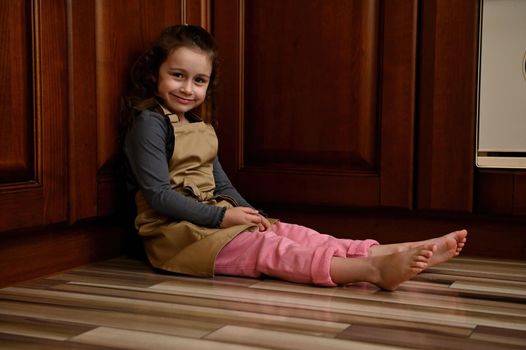 Beautiful preschool age child, a charming European little girl, cute baker confectioner wearing a beige chef's apron, sitting barefoot on the kitchen floor near a cupboard, smiling looking at camera