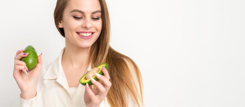 Portrait of a lovely smiling young brunette caucasian woman wearing the white shirt with long hair holding and showing avocado, standing isolated over white background