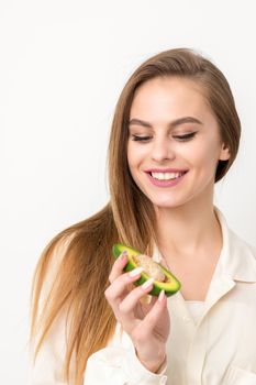 Portrait of a lovely smiling young brunette caucasian woman wearing the white shirt with long hair holding and showing avocado, standing isolated over white background