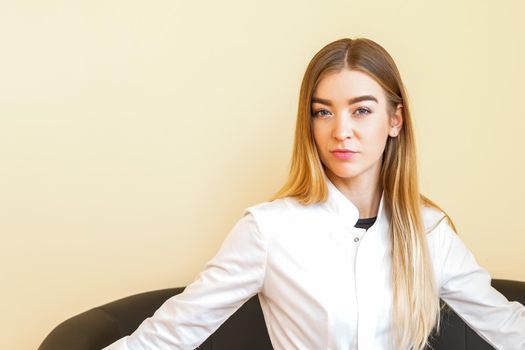 Portrait of a young caucasian doctor with blonde long hair wearing a white coat sitting on the black sofa looking at the camera