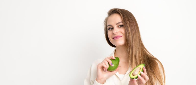 Portrait of a lovely smiling young brunette caucasian woman wearing the white shirt with long hair holding and showing avocado, standing isolated over white background