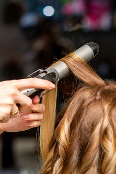 Hairdresser makes curls with a curling iron for the young woman with long brown hair in a beauty salon