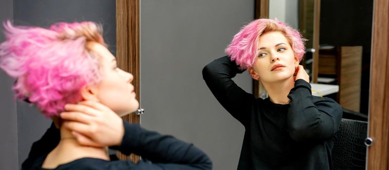 The beautiful young caucasian woman with a new short pink hairstyle looking at her reflection in the mirror checking hairstyle in a hairdresser salon