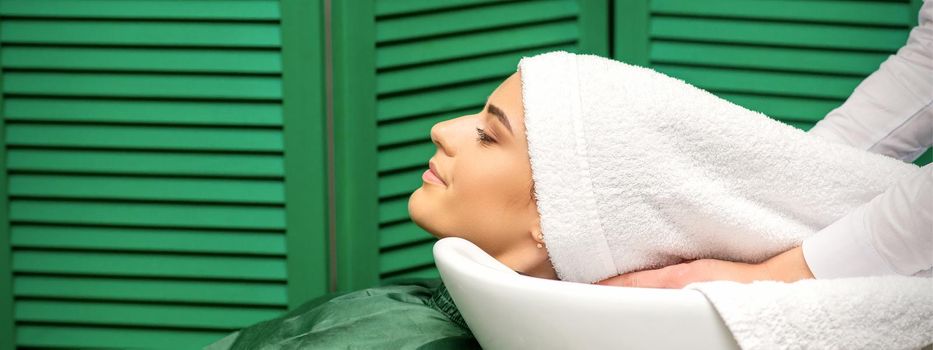 Hairdresser wraps hair of a young caucasian woman in a white towel after washing head in the hairdressing salon