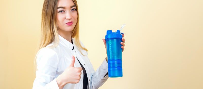 The young Caucasian female doctor wearing a white coat holds a blue plastic bottle with water with thumb up looking at the camera